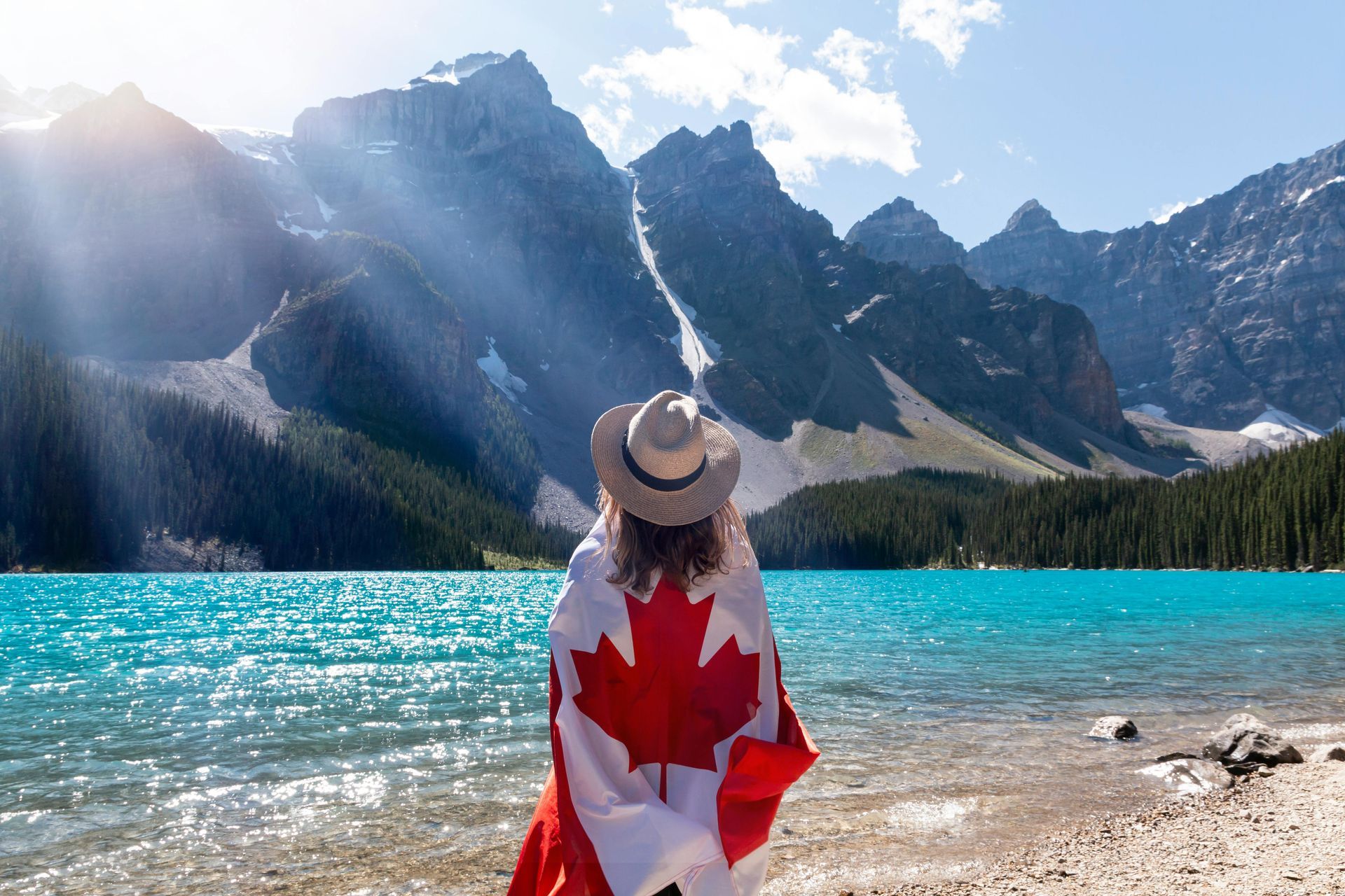 Persona avvolta in una bandiera canadese, di fronte a un lago turchese e alle montagne, sotto un sole splendente.
