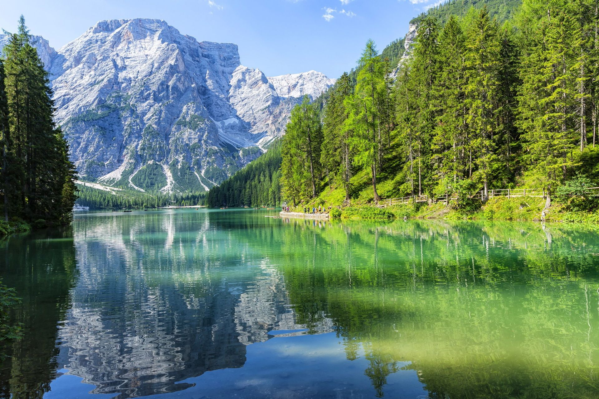 Lago di montagna turchese che riflette la pineta e le cime rocciose frastagliate sotto un cielo azzurro limpido.