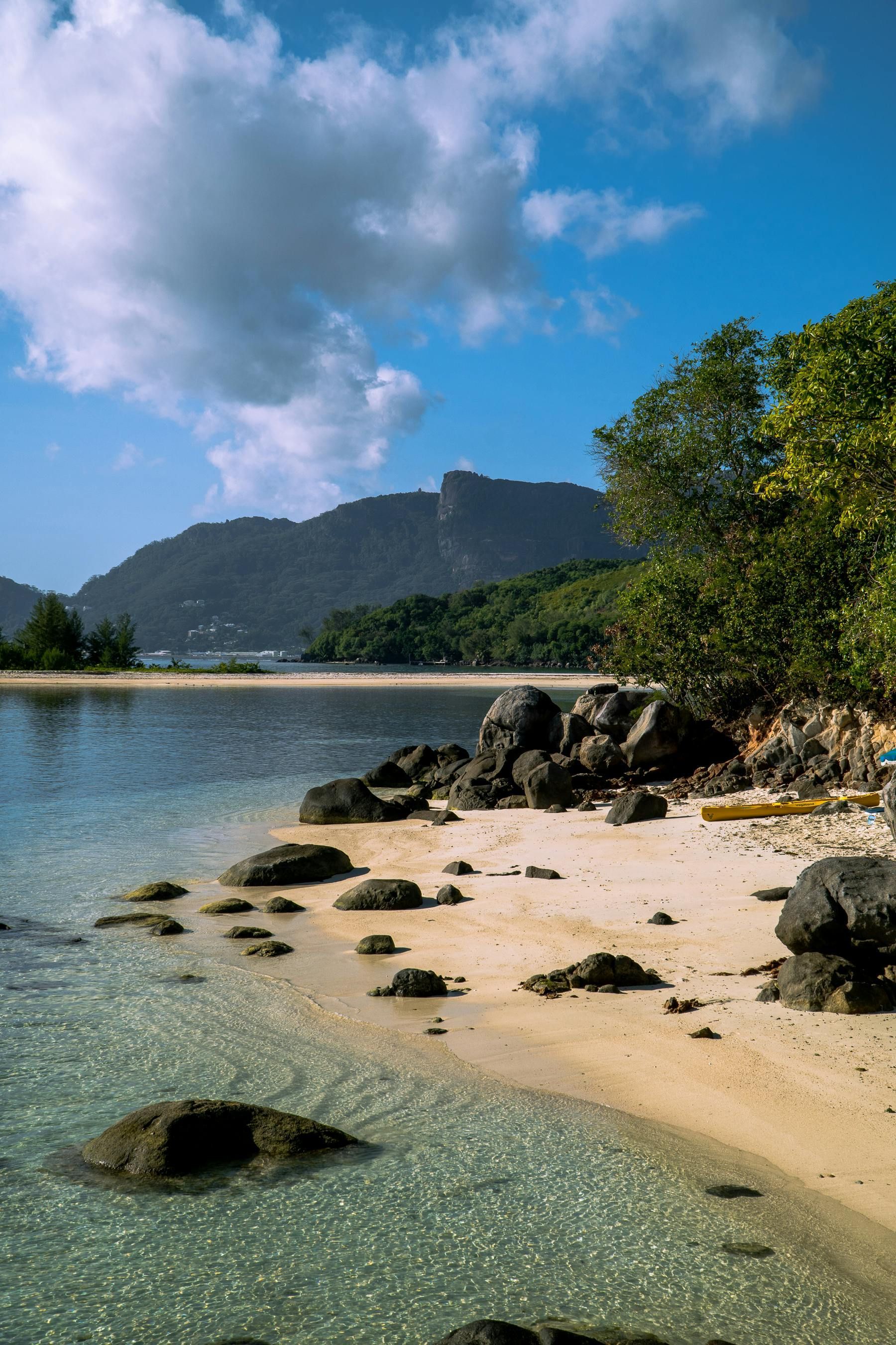 Sandy tropical beach with clear water, rocks, trees, and a distant mountain under a blue sky with clouds