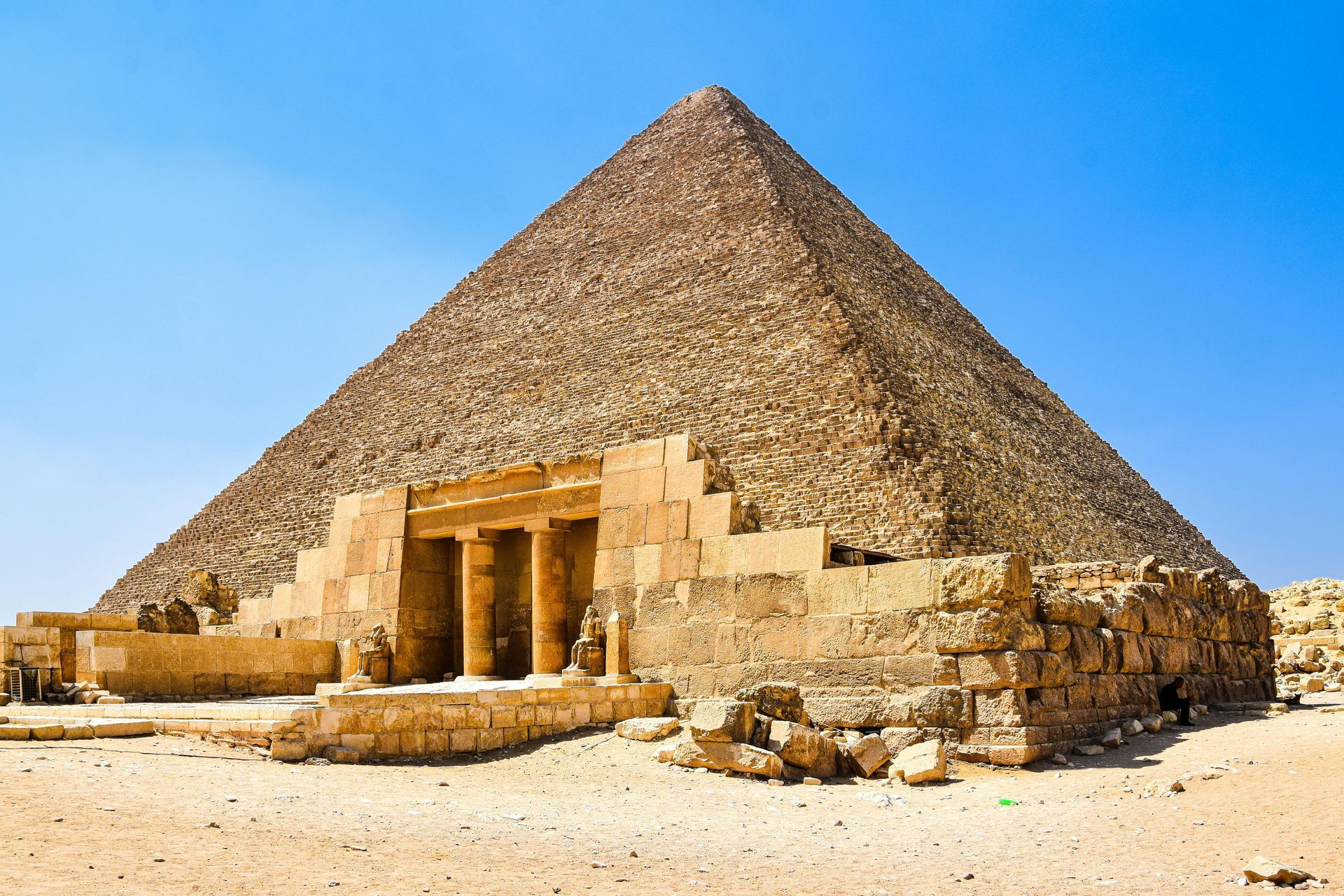 Ancient stone pyramid under a clear blue sky, with a small temple entrance at the base and desert sand around it