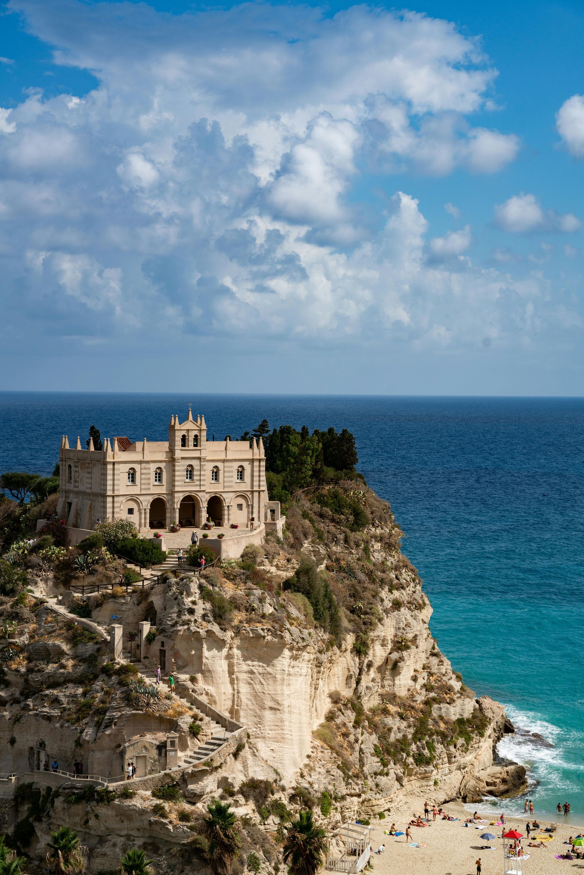 Cliffside castle above turquoise sea and sandy beach under a cloudy blue sky