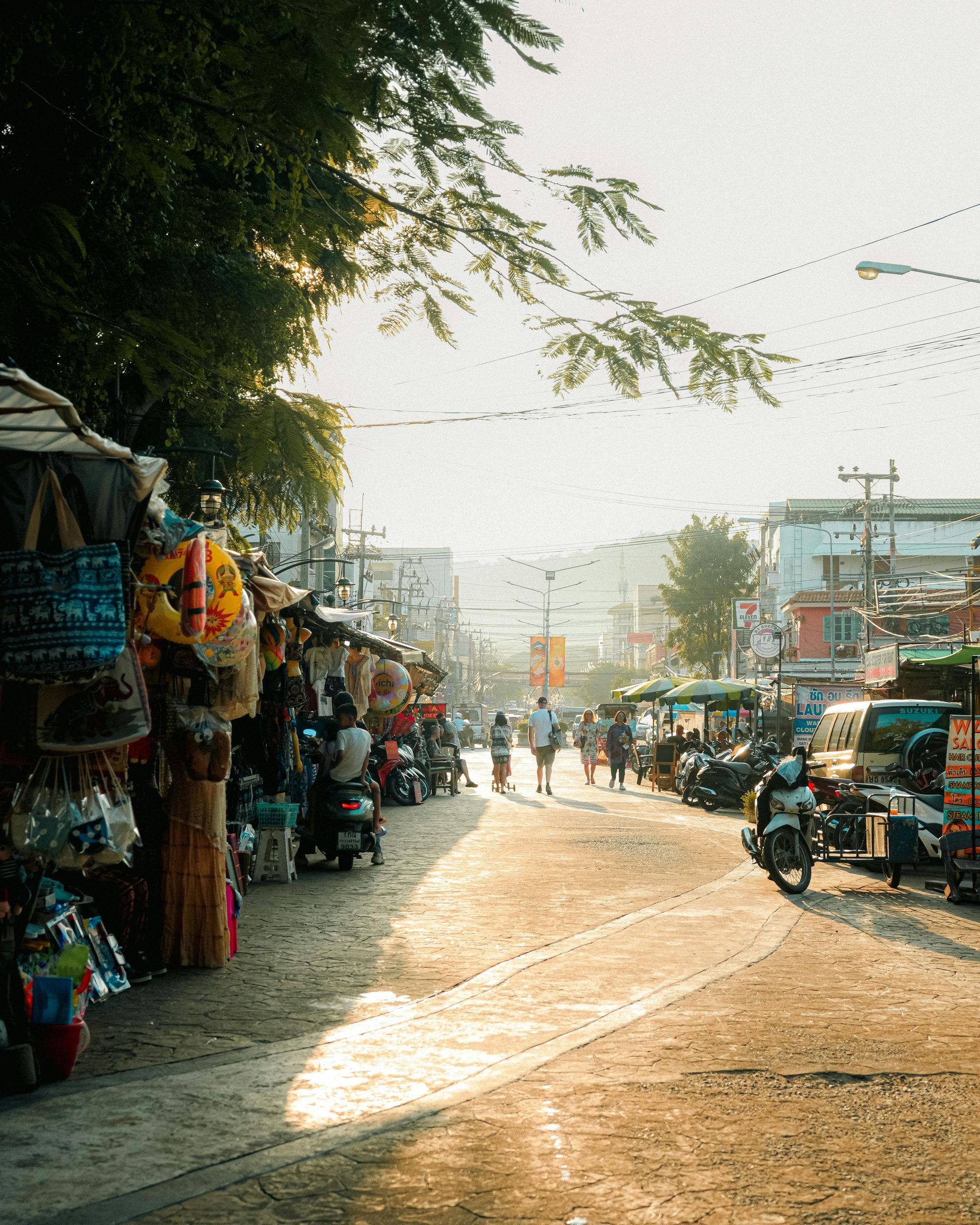 Sunlit street market with stalls, parked motorcycles, and people on a dusty road at dusk