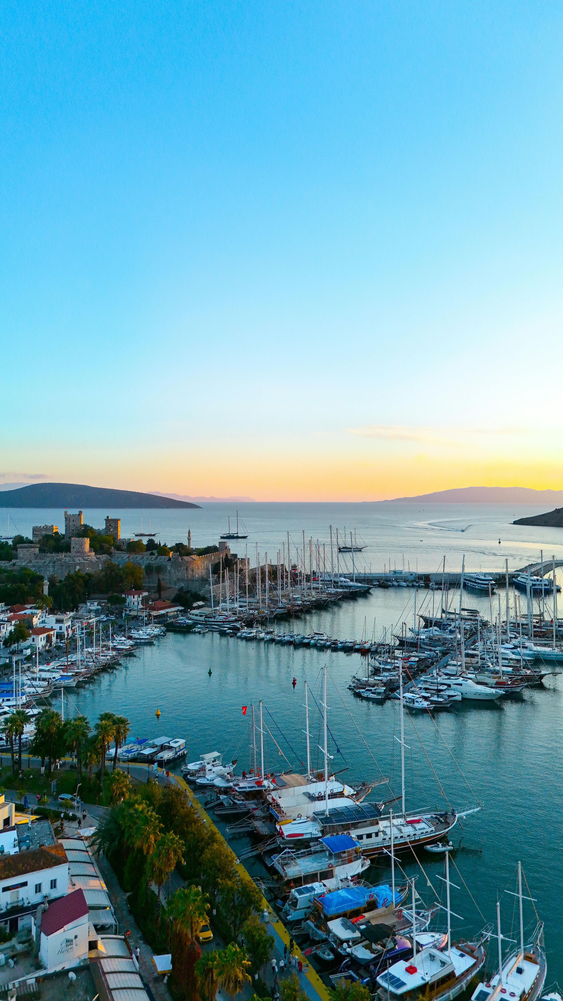 Harbor marina at sunset with boats, calm water, and a pastel sky over distant hills