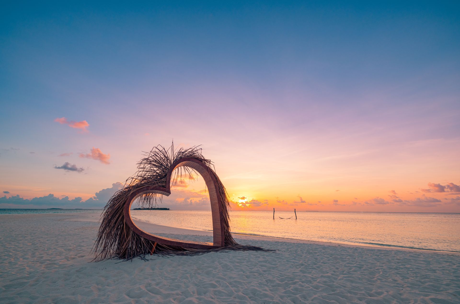 Heart-shaped sculpture on a beach at sunrise, with calm water and a pastel sky.