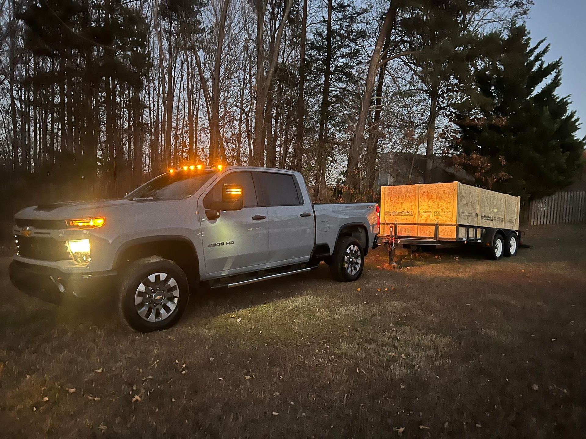 Silver pickup truck towing a lit utility trailer at dusk in a wooded area
