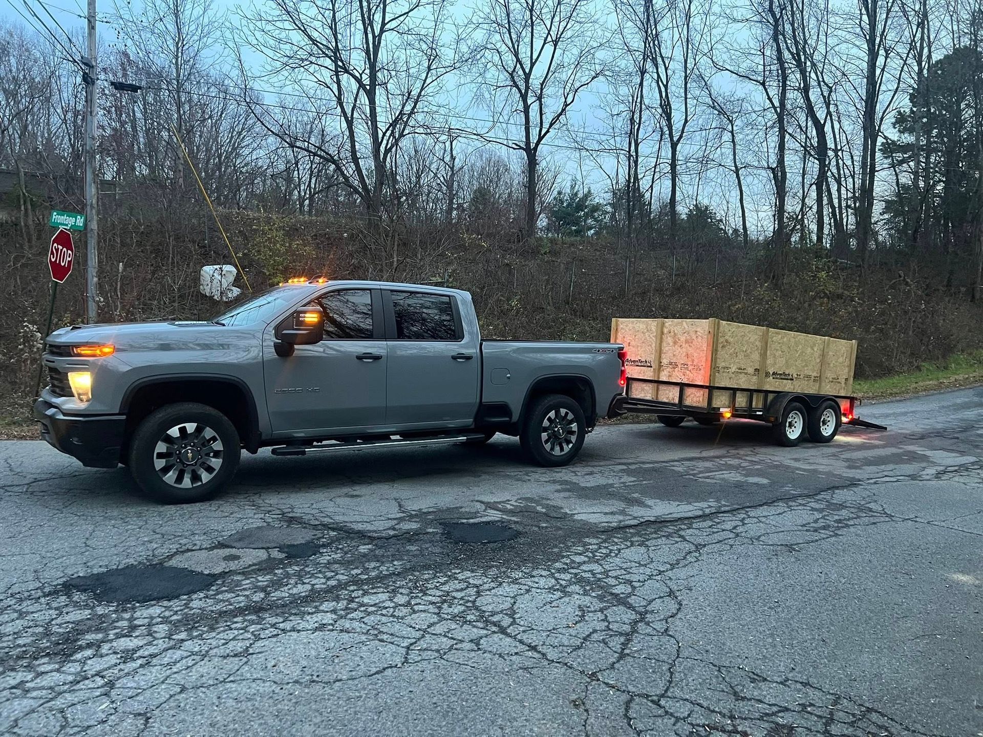 Silver pickup truck towing a small wood-sided trailer on a wet road beside leafless trees.