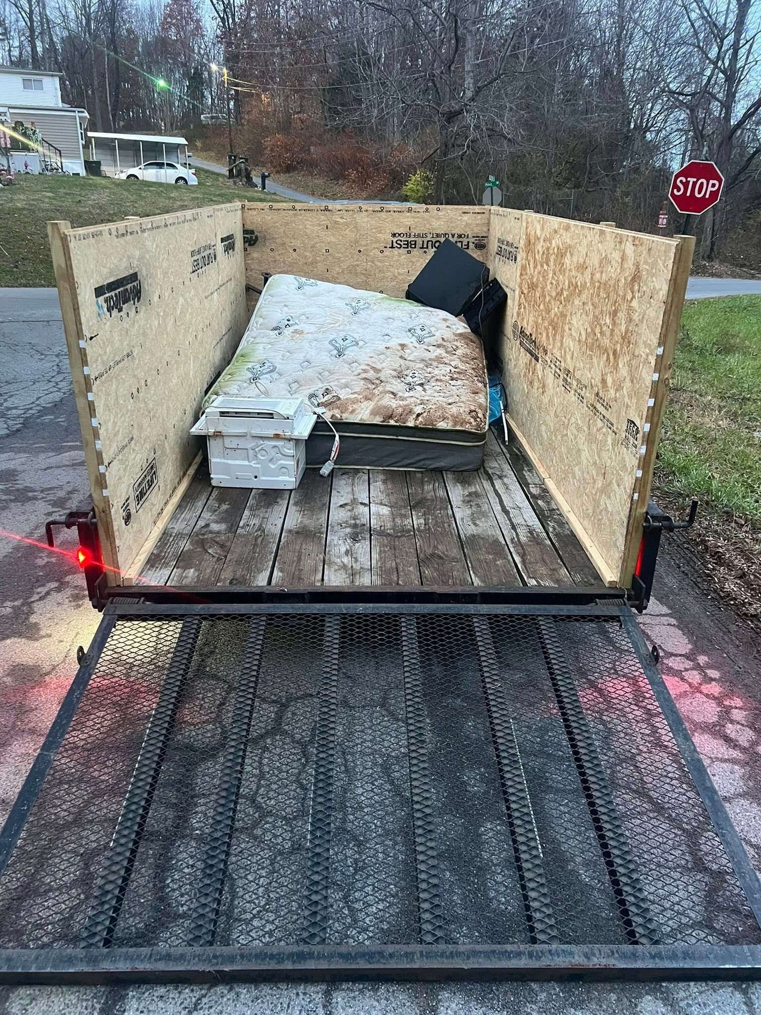 Open utility trailer with plywood sides and a covered load on a wet road, viewed from behind.