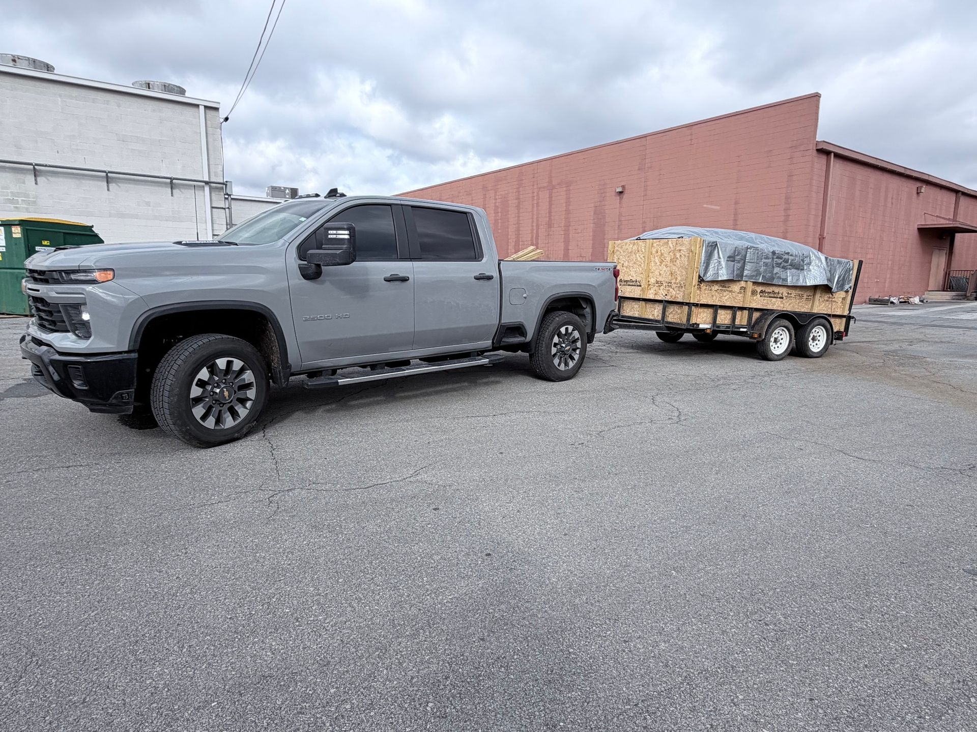 Gray pickup truck towing a wood-loaded trailer in an industrial lot under cloudy skies