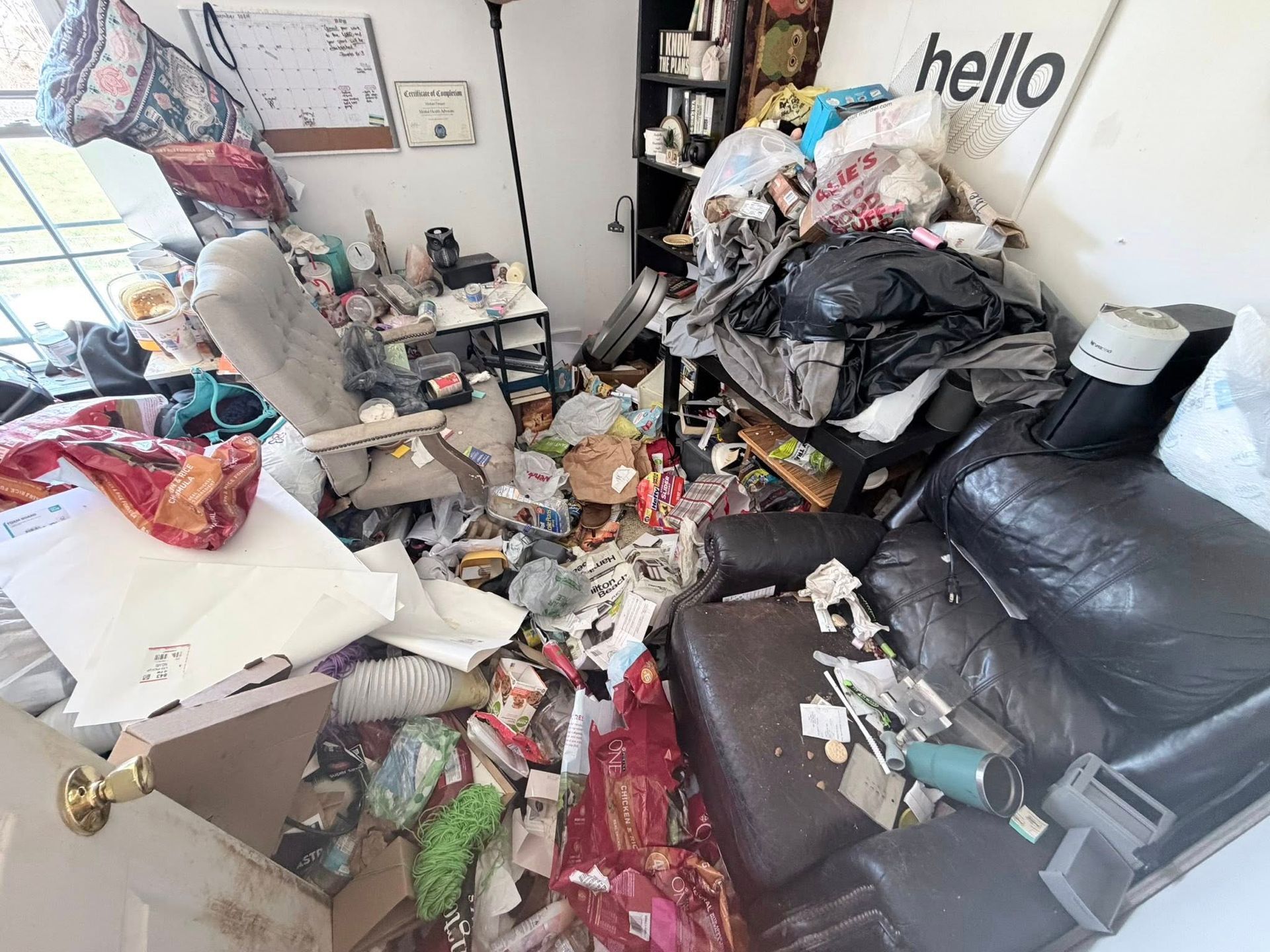 Cluttered living room with piles of trash, papers, and bags on the floor and a black couch on the right
