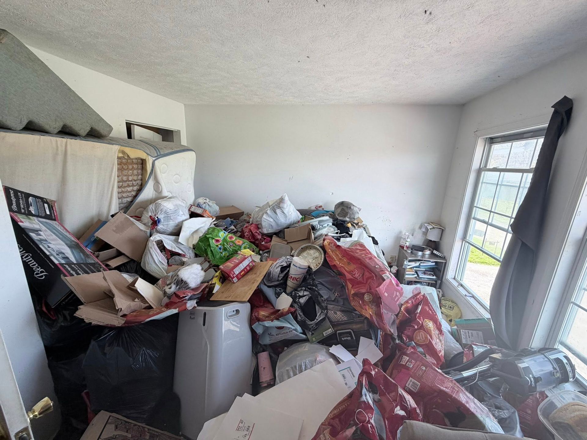 Cluttered room piled high with bags, boxes, and household items near a window and sliding glass door