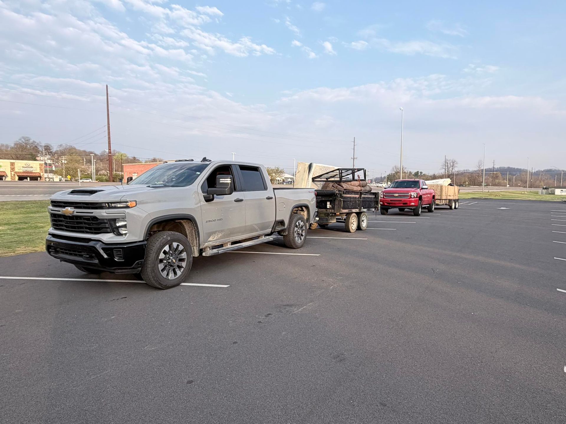 Silver pickup truck towing a trailer and red car on an open parking lot road under a cloudy sky