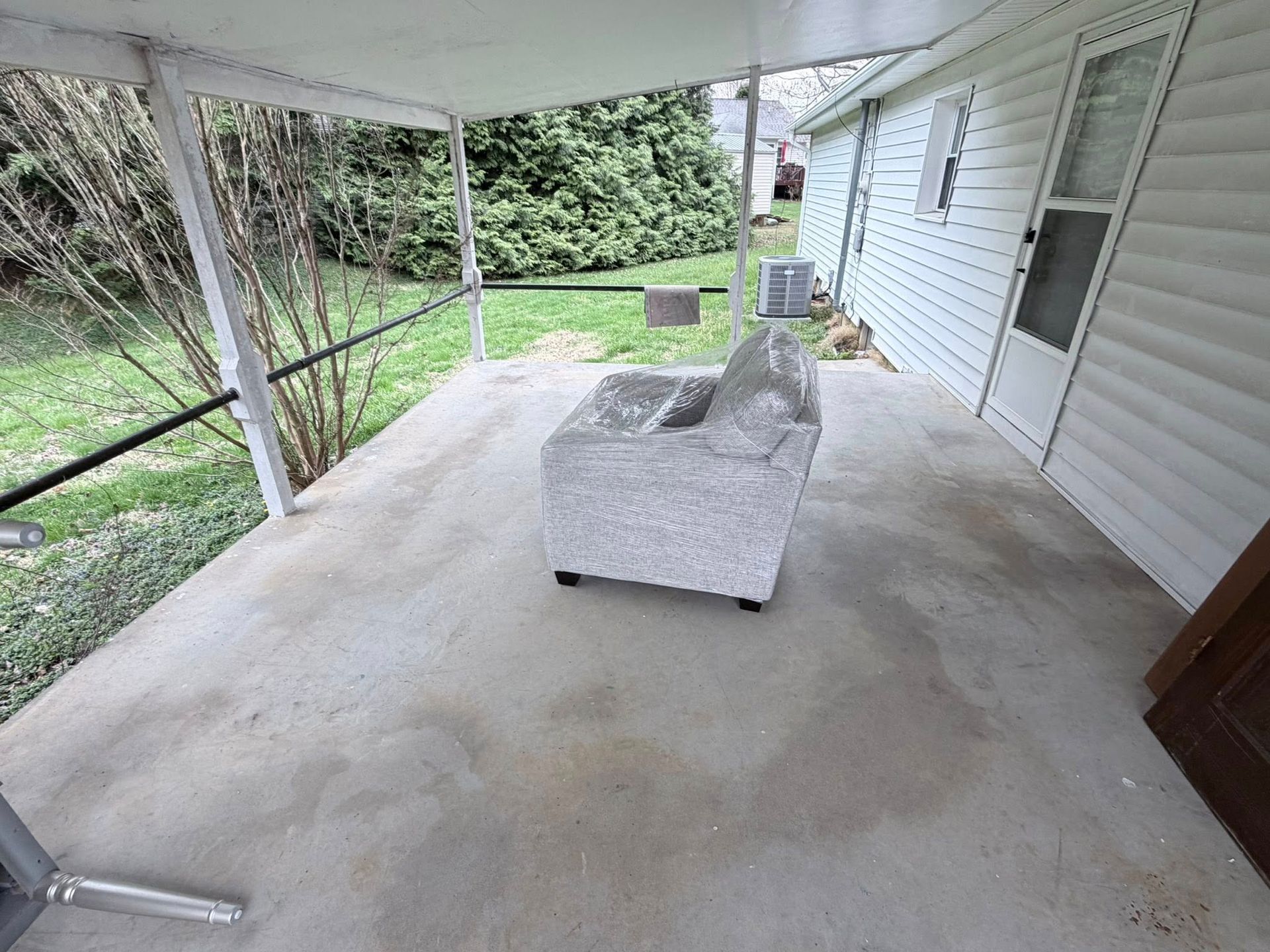 Covered concrete patio with a gray sofa, white siding, and a view of the yard and garden hose