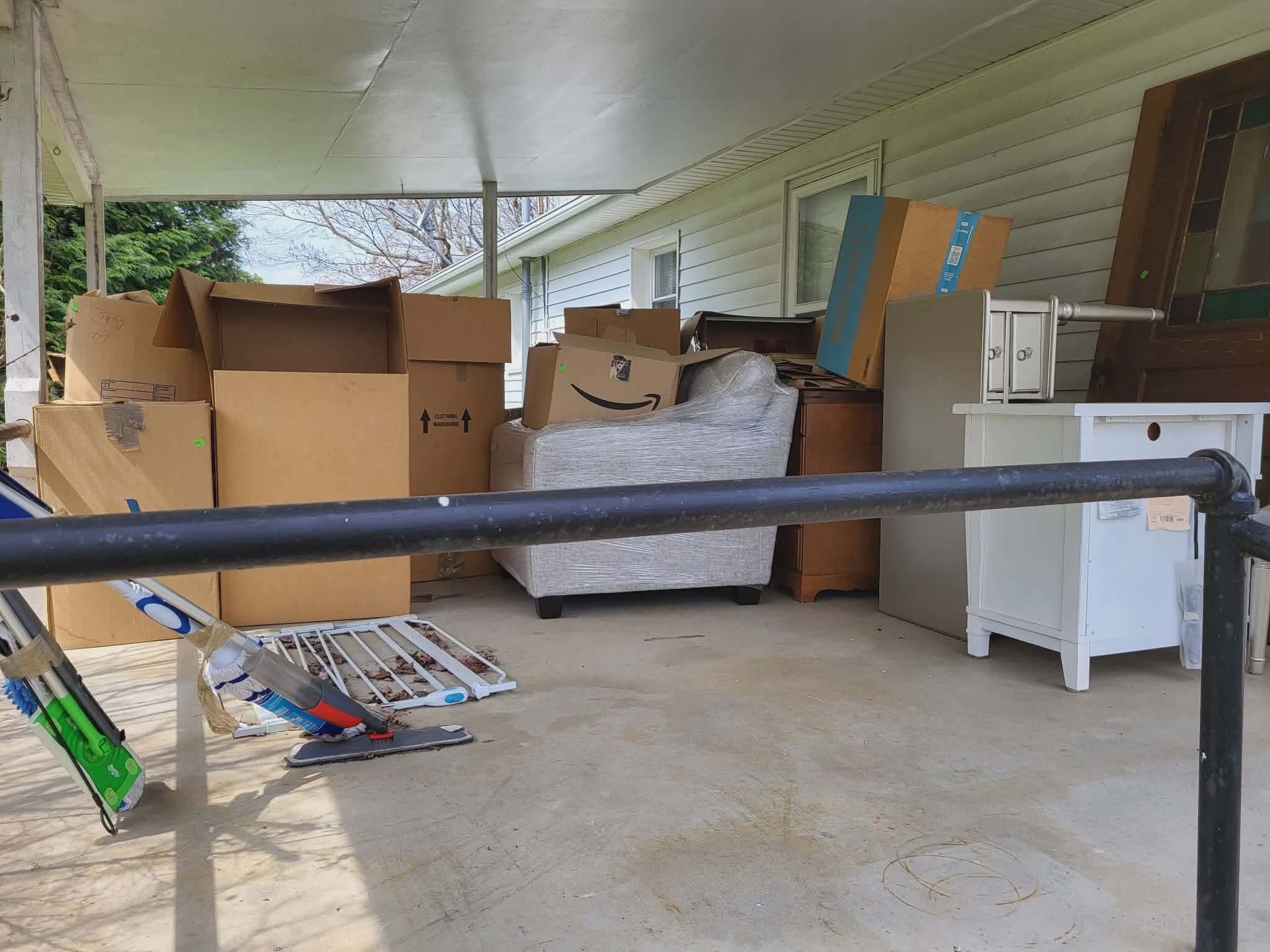 Cluttered porch with cardboard boxes, furniture, and household items under a covered roof