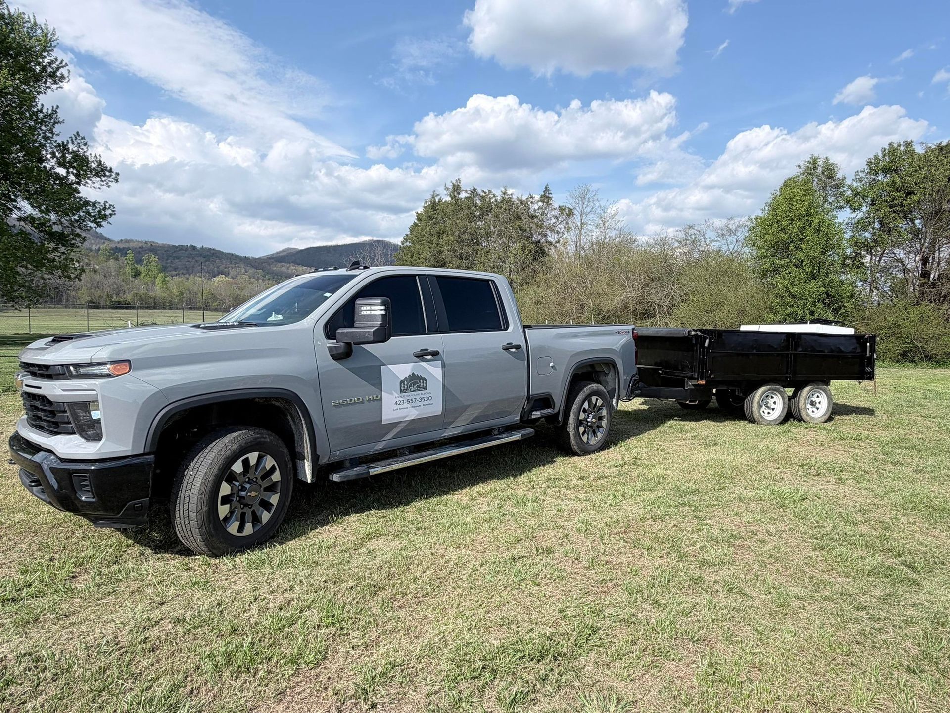 Silver pickup truck parked on grass, towing a black trailer under a partly cloudy sky.