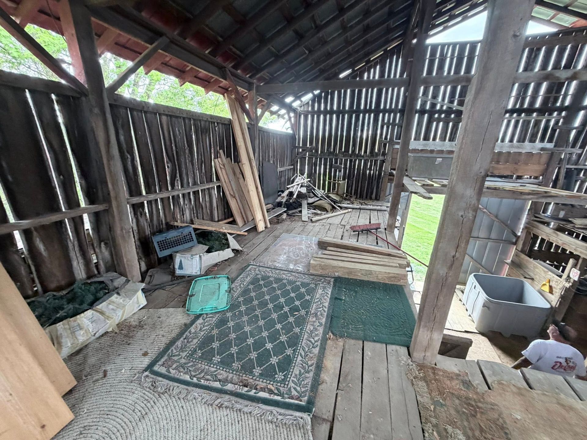 Interior of a rustic barn with worn wooden walls, scattered mats, and household items on the floor.