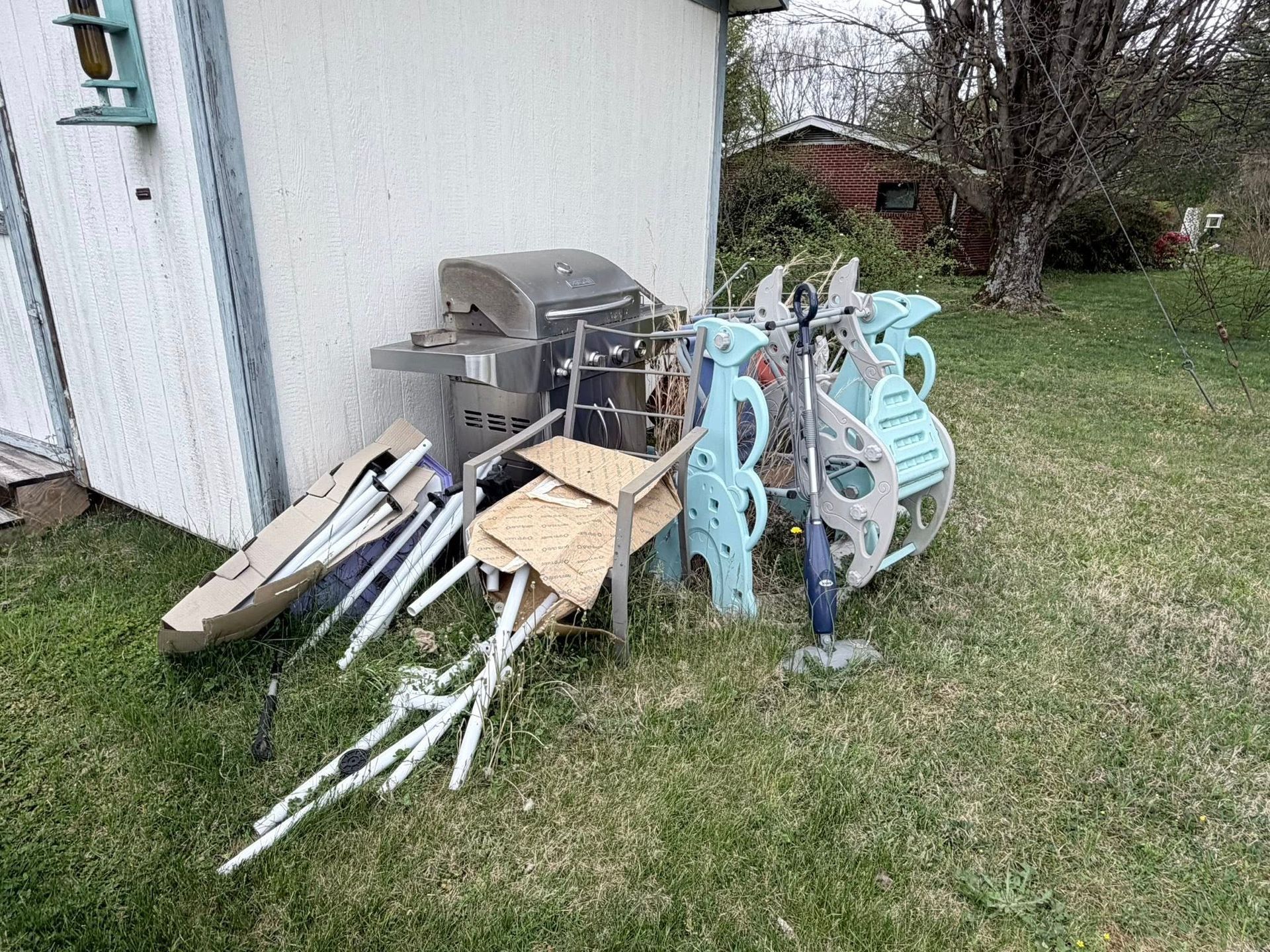 Pile of outdoor tools and scrap leaning against a white building on a grassy yard.