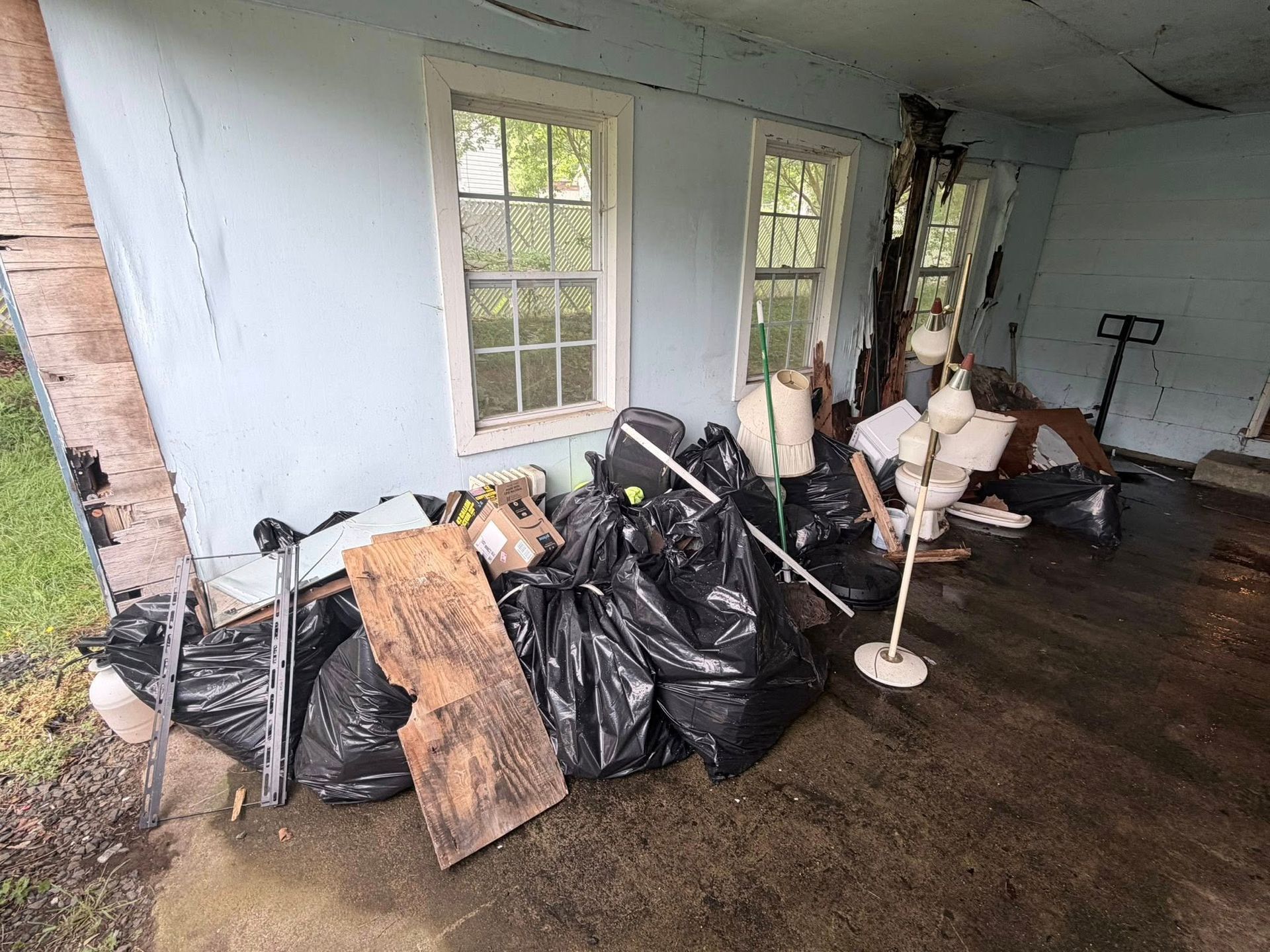 Cluttered porch with black trash bags, broken boards, and debris beside a white exterior wall and windows