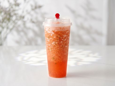 A tall orange slush drink in a clear cup with a red cherry lid, on a bright white table