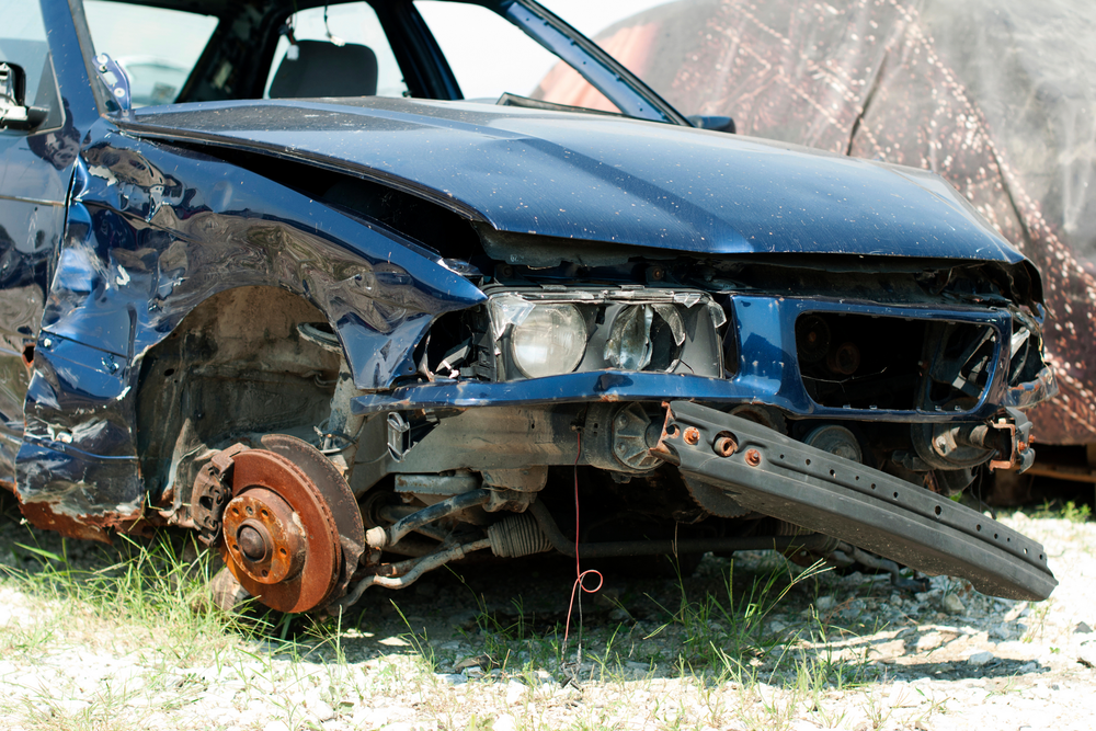 Auto blu gravemente danneggiata, con la parte anteriore schiacciata e una ruota mancante, sull'erba a bordo strada.