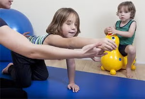 Toddler on blue mat reaching for yellow toy while another child sits nearby in a playroom