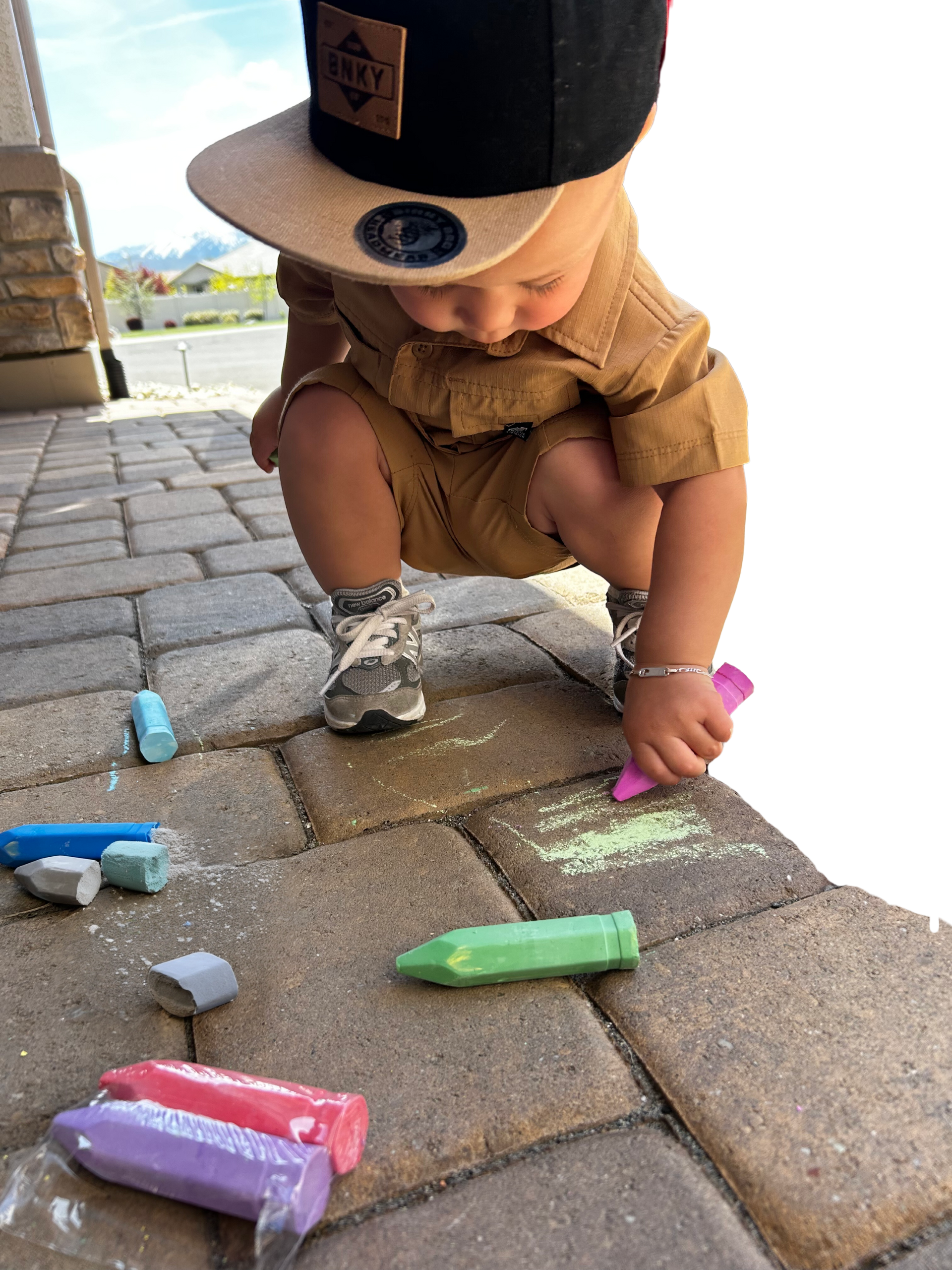 Image of a young boy aged 1 year squatting down drawing on driveway pavers with a pink piece of large chalk 