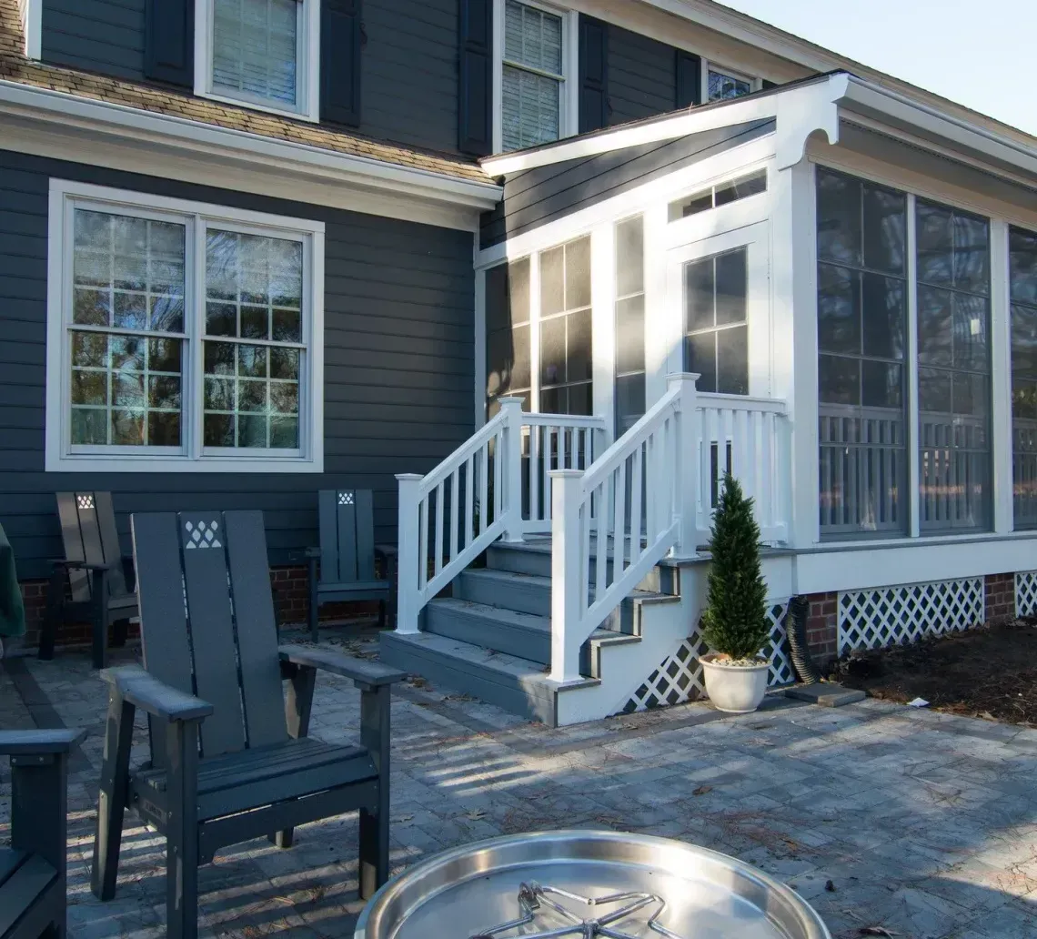Backyard view of a light gray house with a porch, windows, and an enclosed sunroom over dry grass.