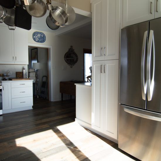 Modern kitchen with stainless steel fridge, white cabinets, hanging pots, and sunlit hardwood floor