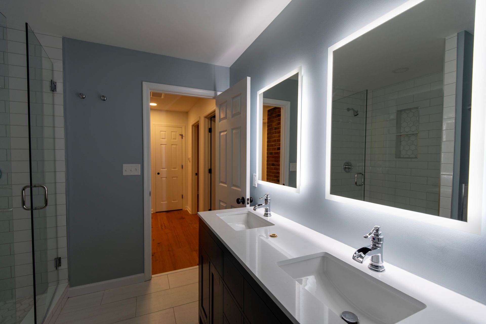 Modern bathroom with double sinks, lit mirrors, gray walls, and a doorway to a hallway