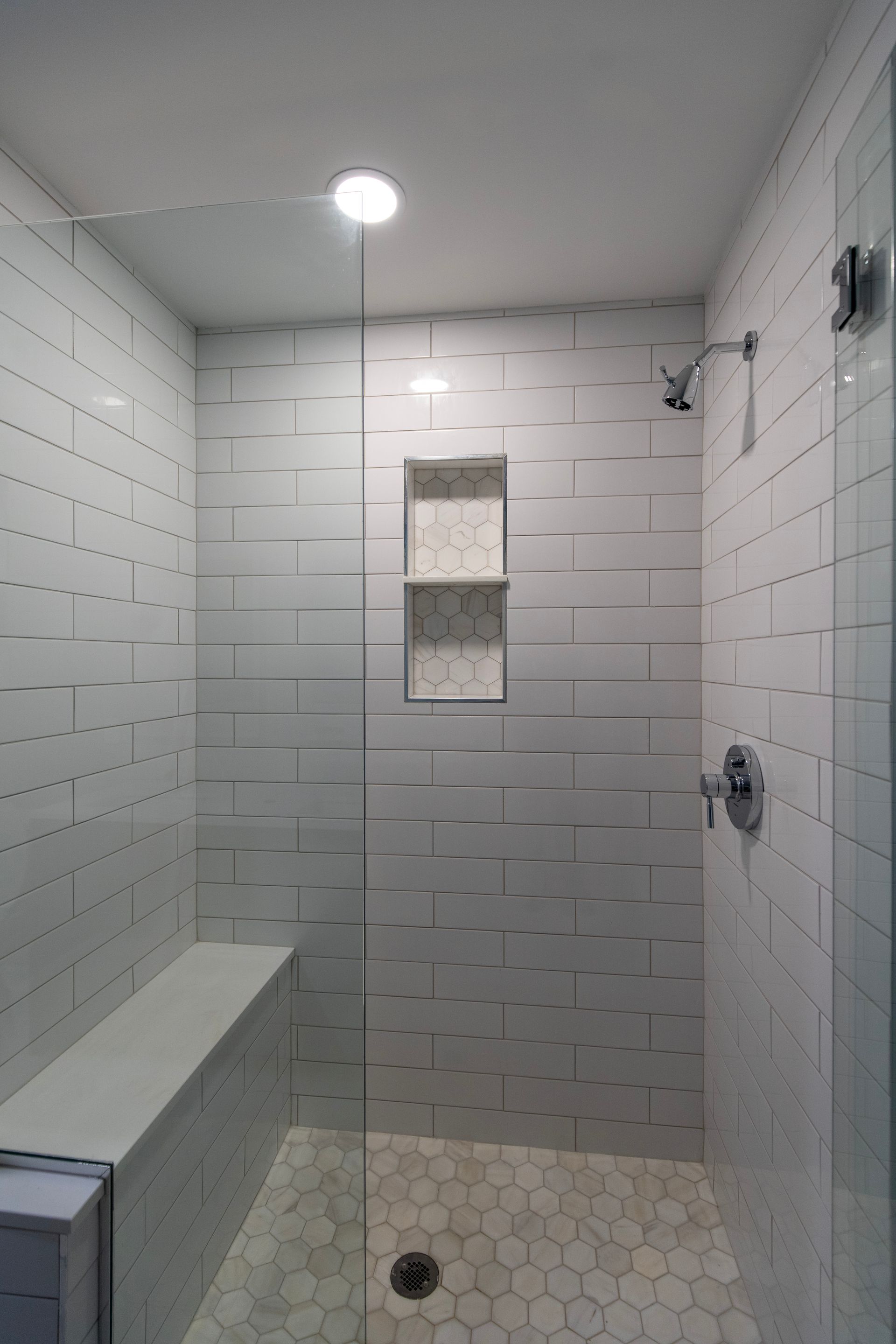 Tiled walk-in shower with a bench, hexagon floor, central drain, and recessed shelf under a ceiling light