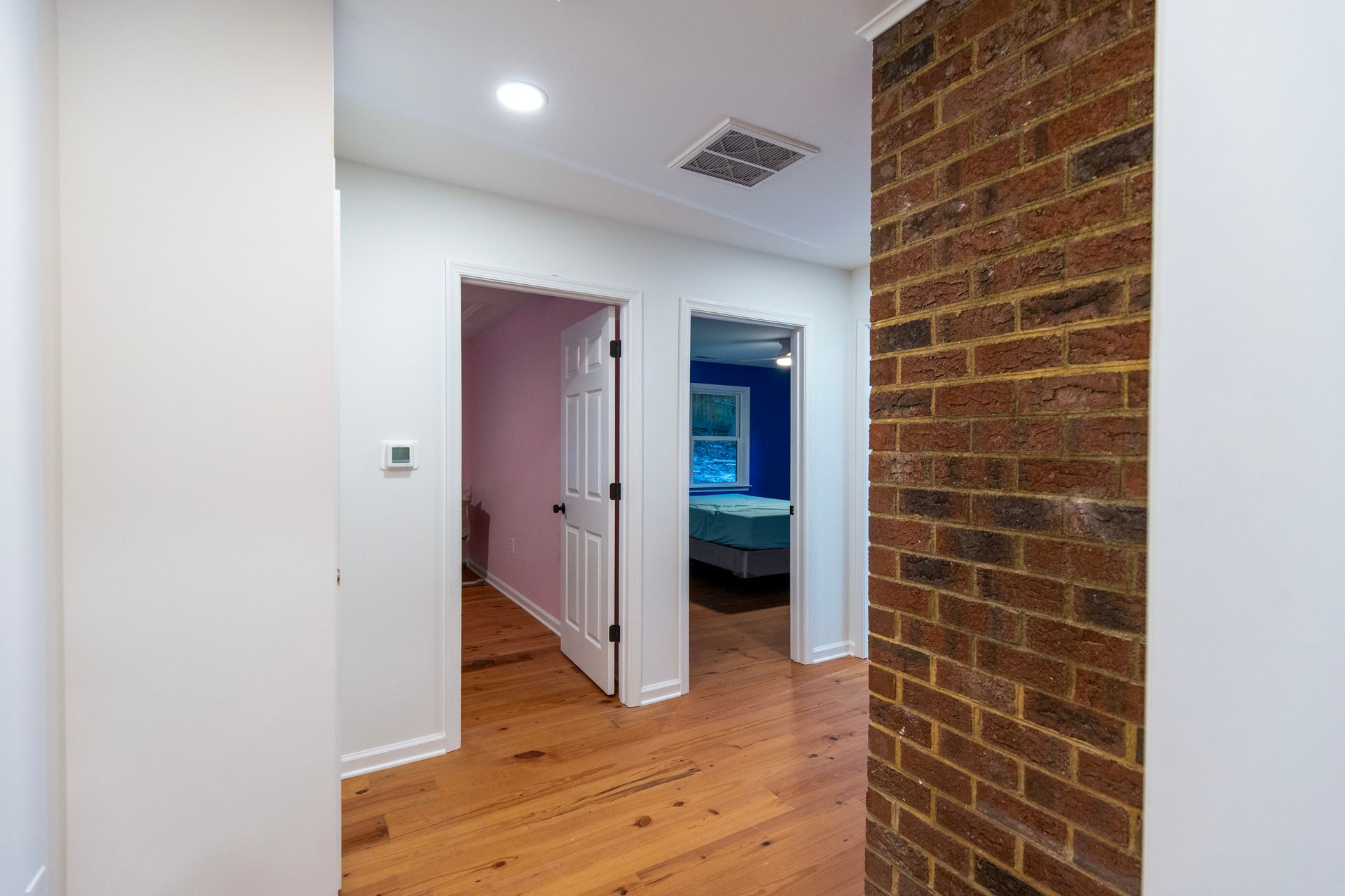 Hallway with white walls, wood floor, brick accent wall, and open doorways to a bedroom and lit room