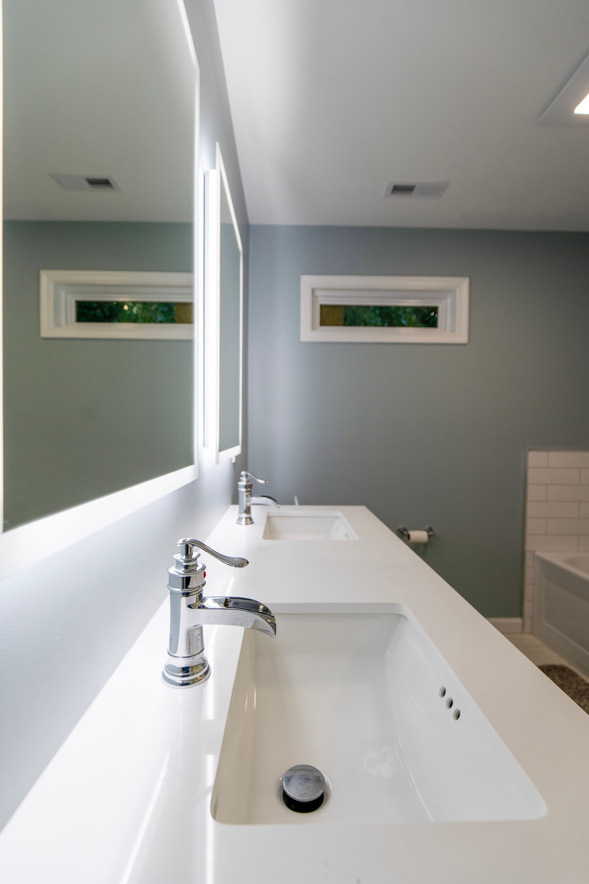 Modern bathroom with a white sink and countertop, mirror, gray walls, and small high window.