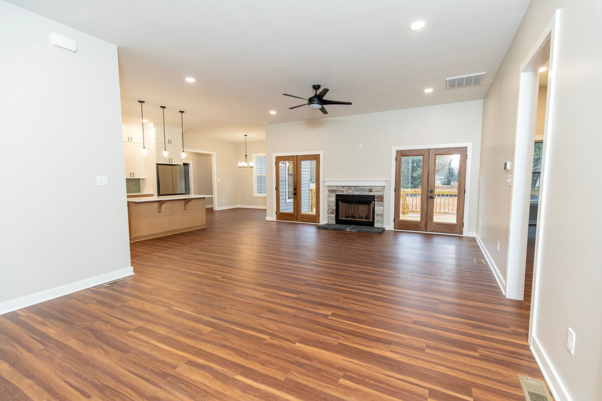 Spacious empty living room with wood floors, fireplace, ceiling fan, and kitchen bar in the background