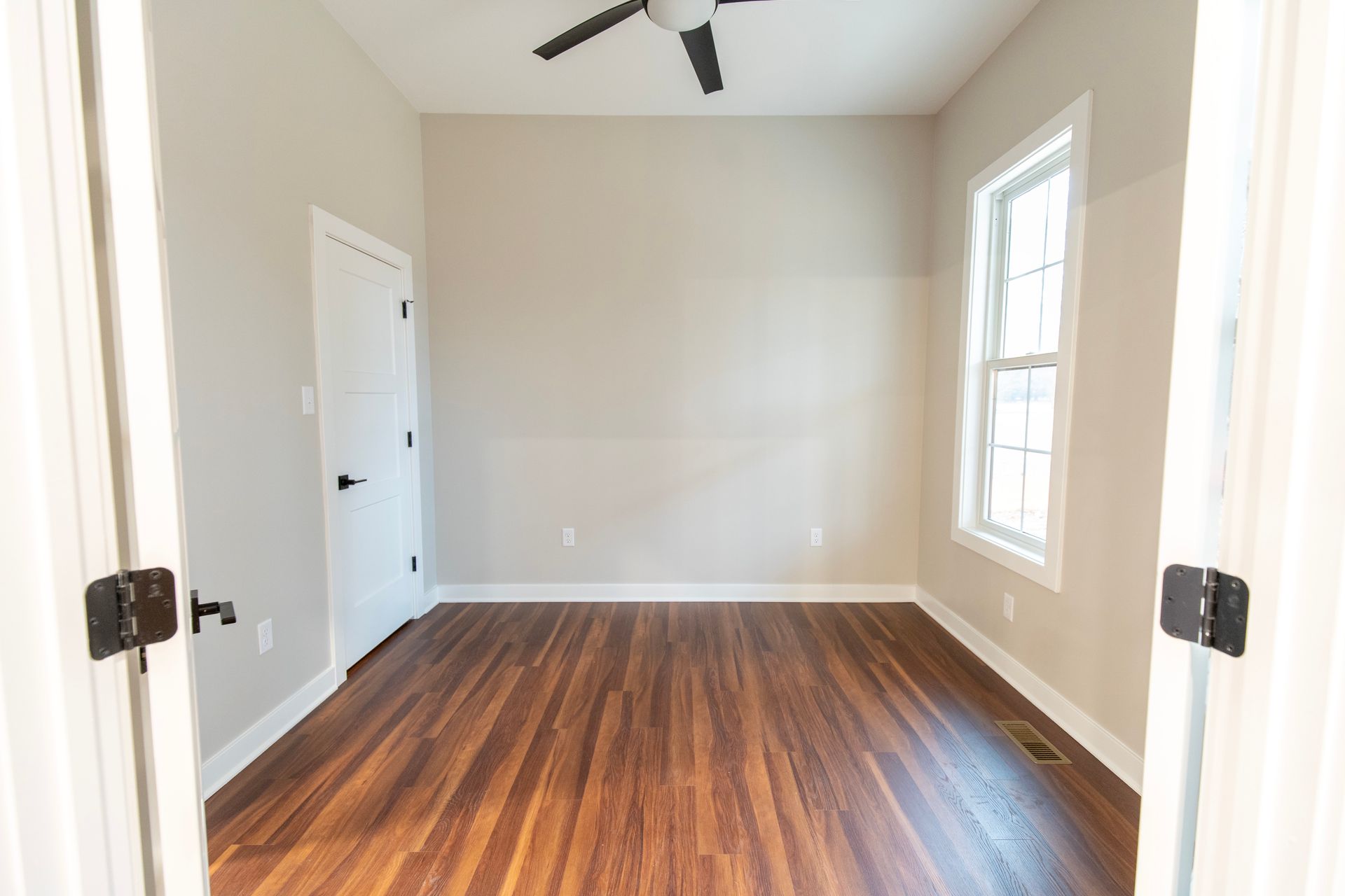 Empty beige room with dark wood floors, ceiling fan, and two windows by a white door