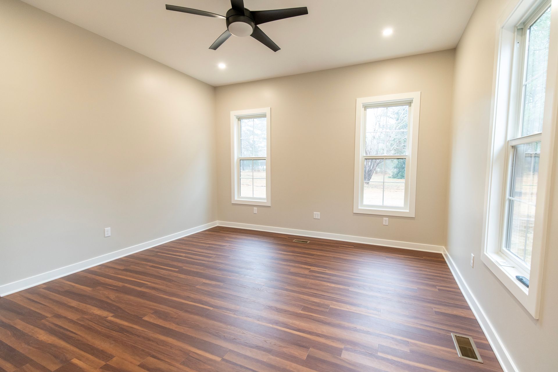 Empty beige room with wood floor, ceiling fan, and three windows.
