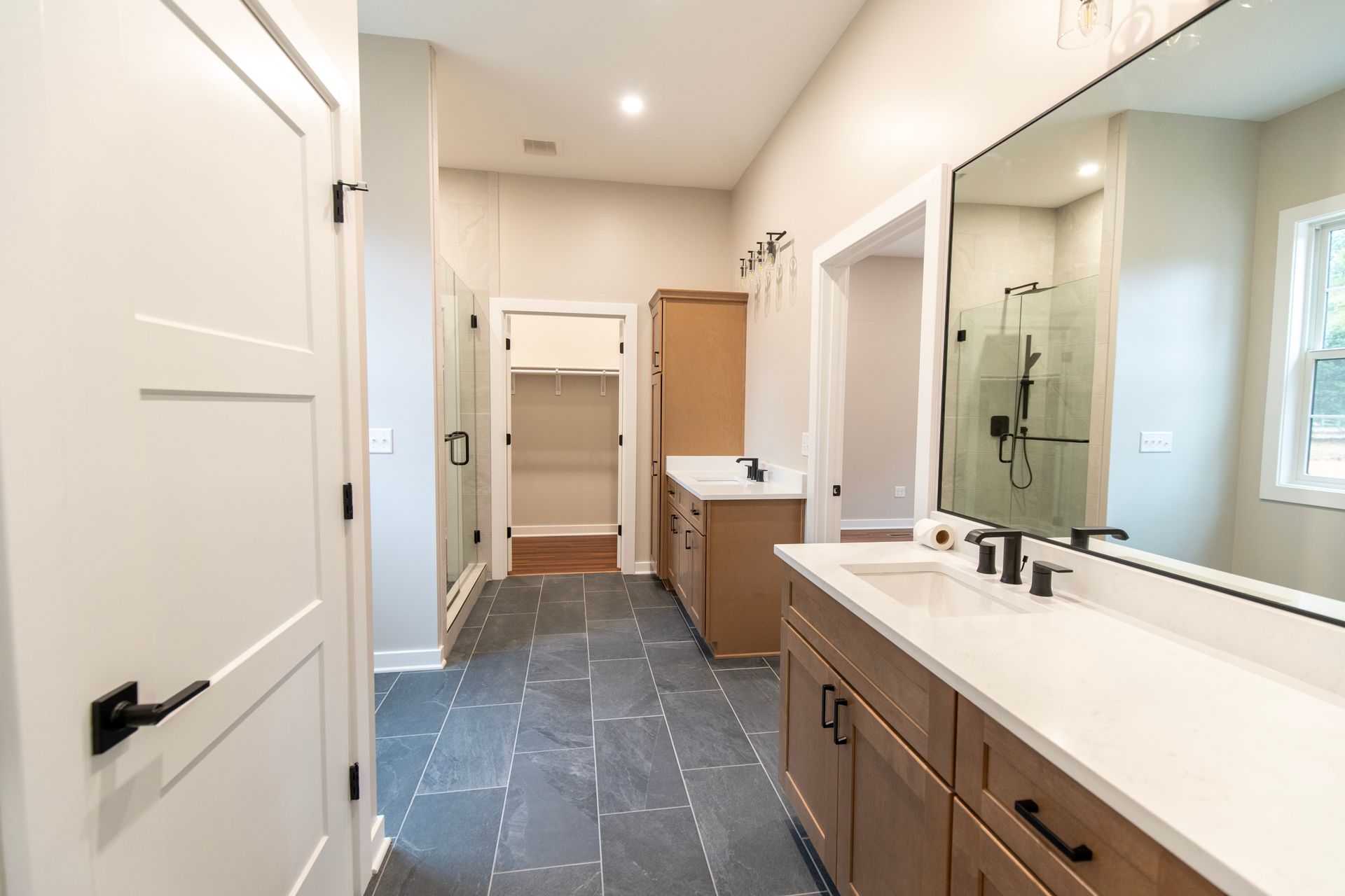 Bright modern bathroom with white walls, dark tile floor, double vanity, mirror, shower, and open closet area