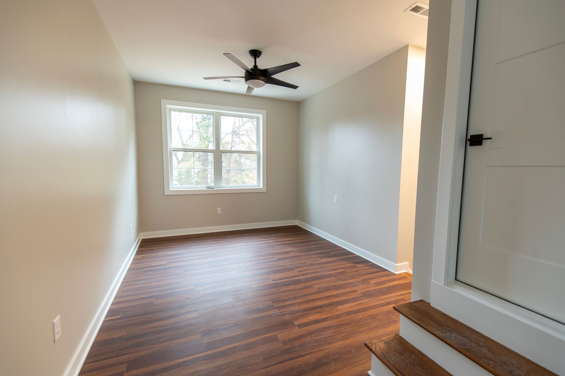 Empty room with wood floors, white walls, window, and ceiling fan, viewed from the doorway