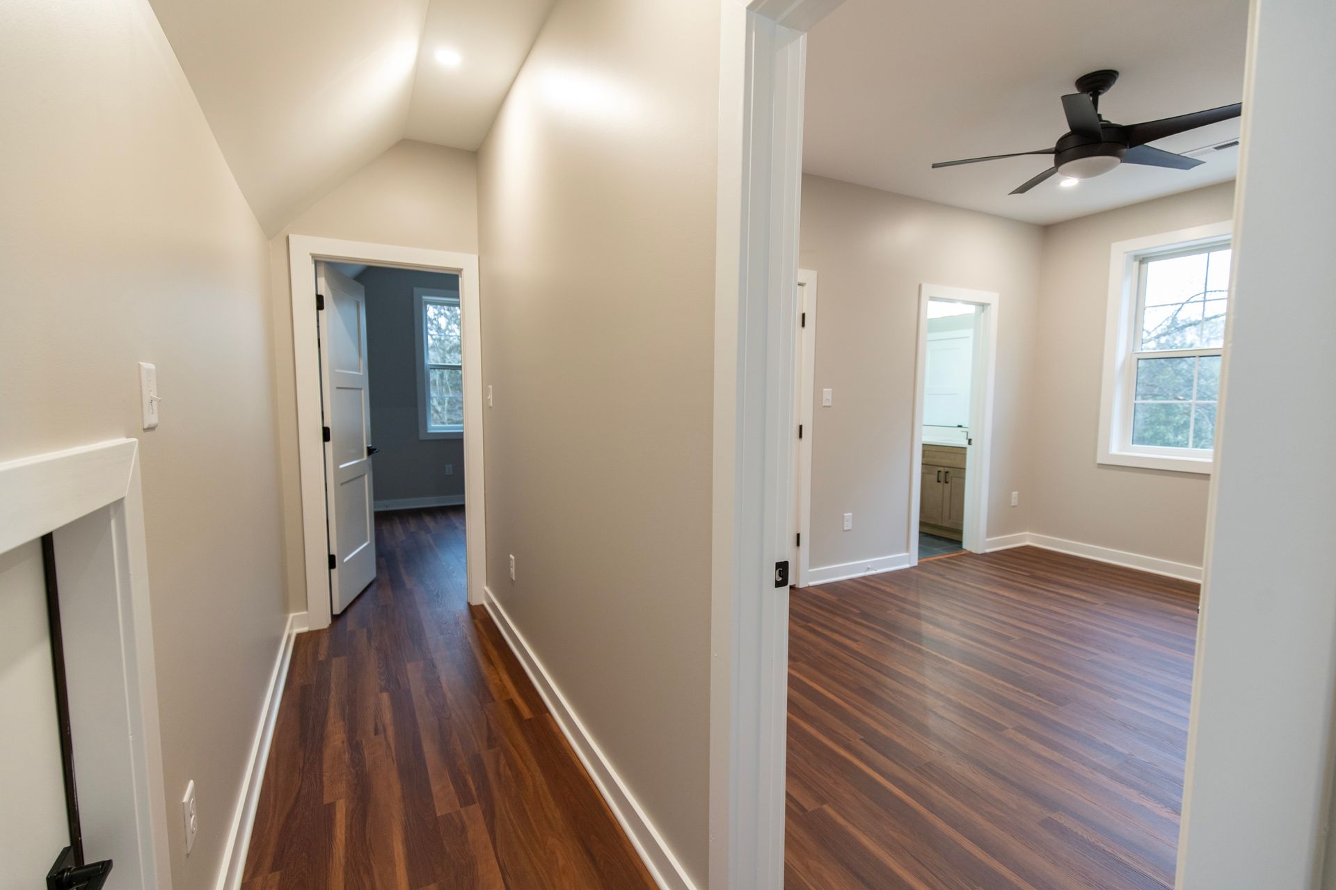 Narrow hallway with dark wood floors leading to a bright room with a ceiling fan and window.