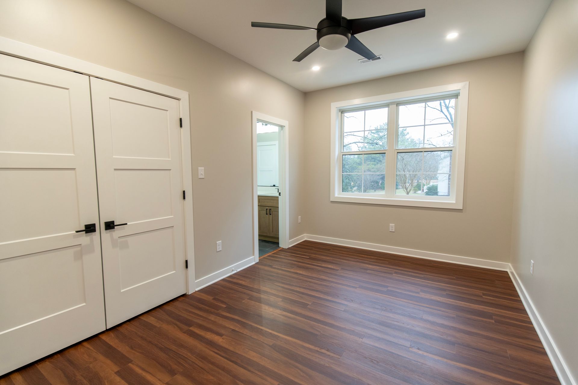 Empty beige bedroom with dark wood floors, white closet doors, ceiling fan, and a window.