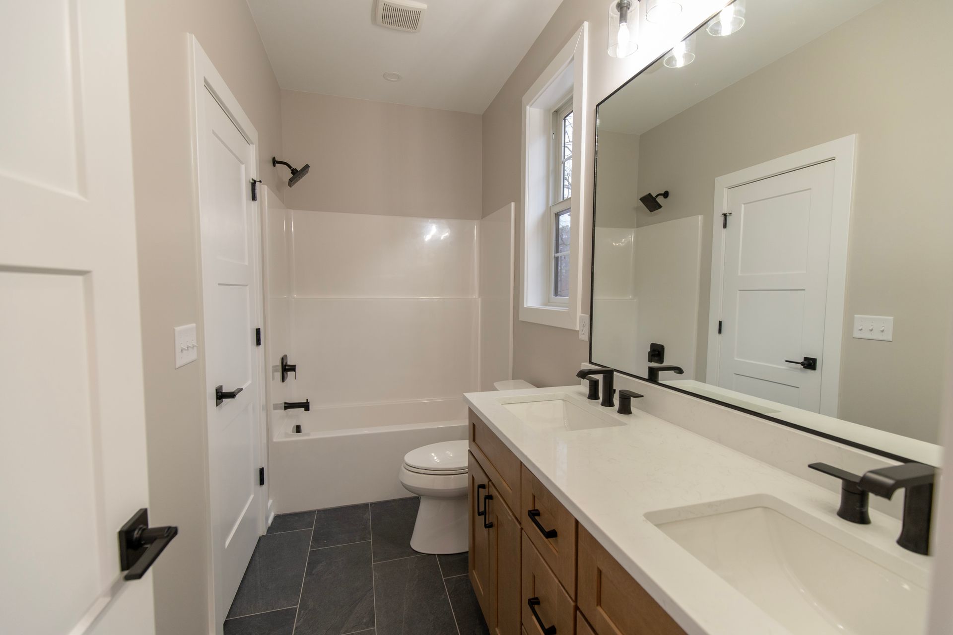 Bright modern bathroom with double vanity, large mirror, toilet, tub, and black fixtures.