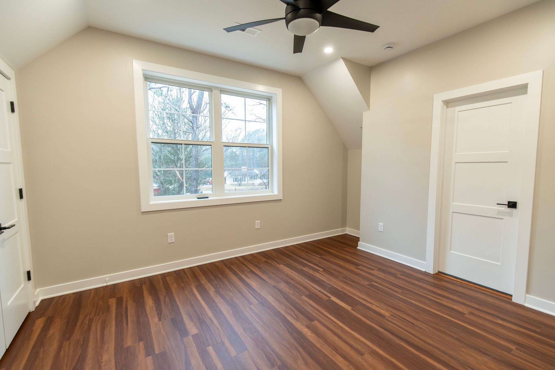 Empty beige bedroom with wood floors, a window, ceiling fan, and white closet doors