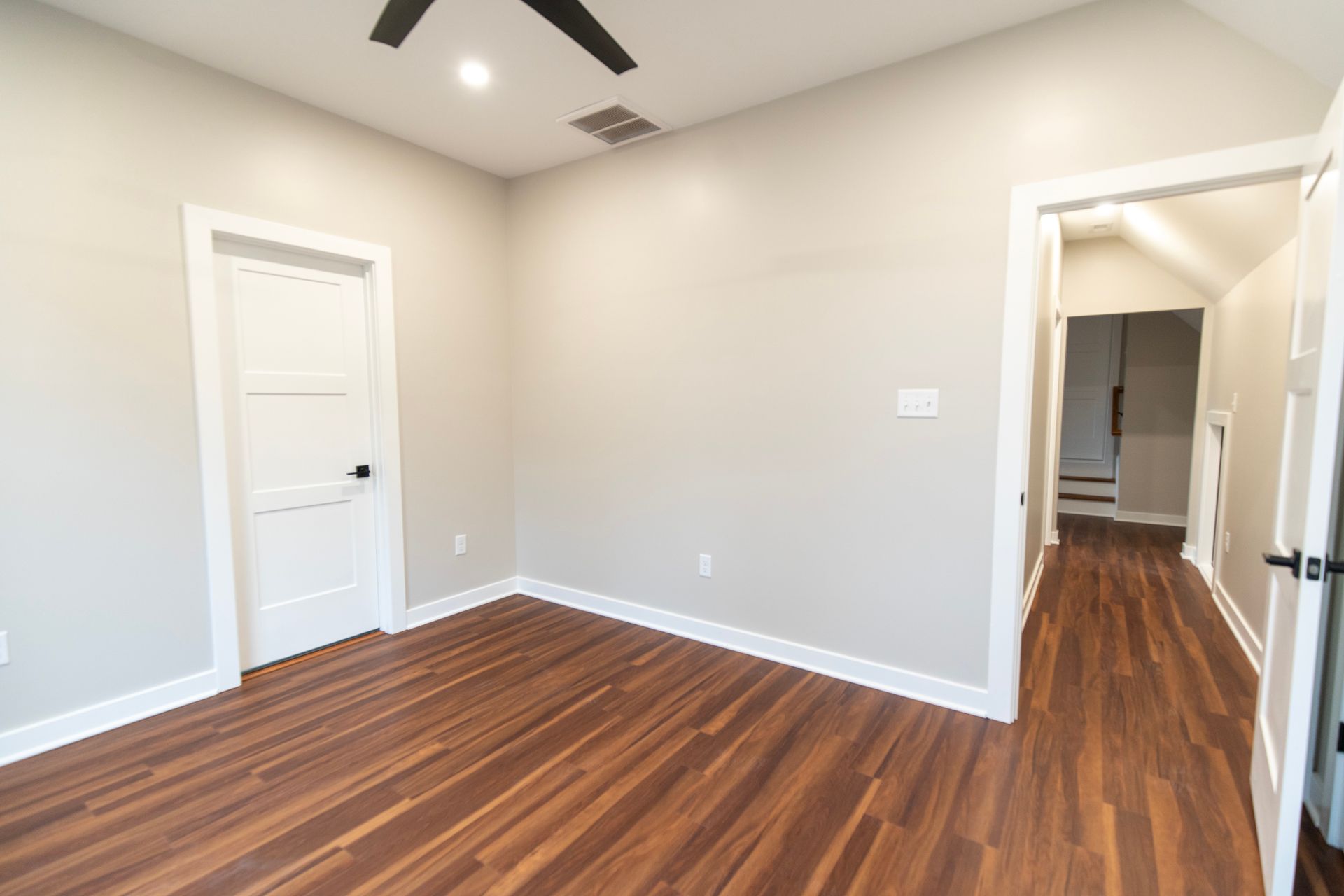 Empty beige room with wood floors, white doors, and a hallway opening to the right
