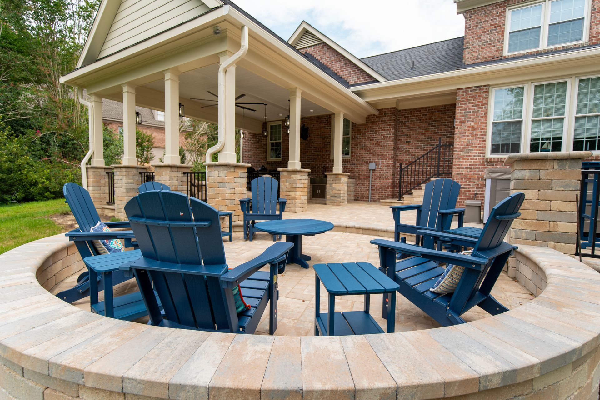 Blue Adirondack chairs around a circular stone fire pit on a brick patio with a covered porch behind.