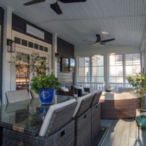 Covered porch with wicker dining chairs, glass table, potted plants, and ceiling fan in bright sunlight