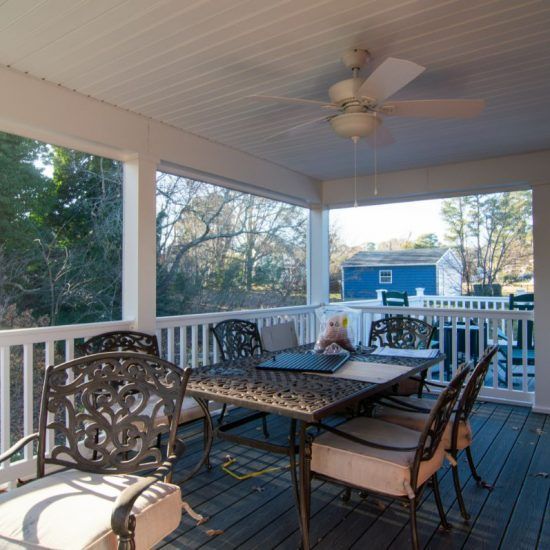 Covered porch with white railing, ceiling fan, and metal patio table and chairs overlooking a blue building.