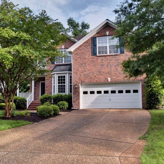 Brick suburban house with a white garage, curved driveway, and green trees framing the front yard