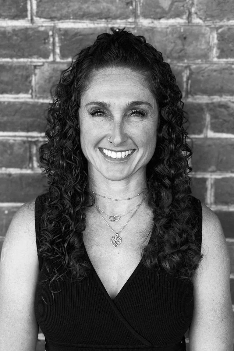 Woman smiling in a black top, standing before a brick wall.