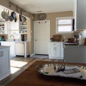 Bright kitchen with white cabinets, hanging pans, a door, and a wooden table in the foreground.