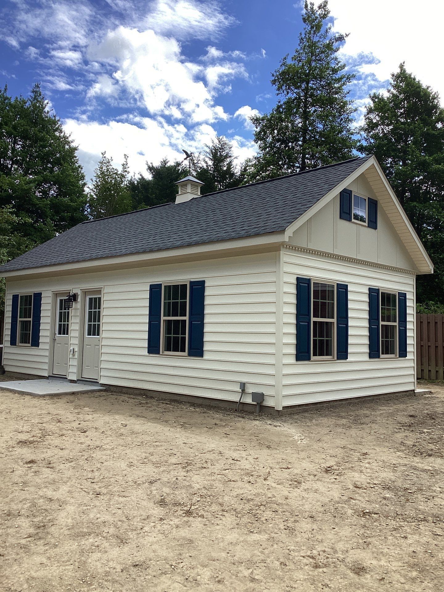 Small beige house with blue shutters and a dark roof, set on a sandy lot under a partly cloudy sky