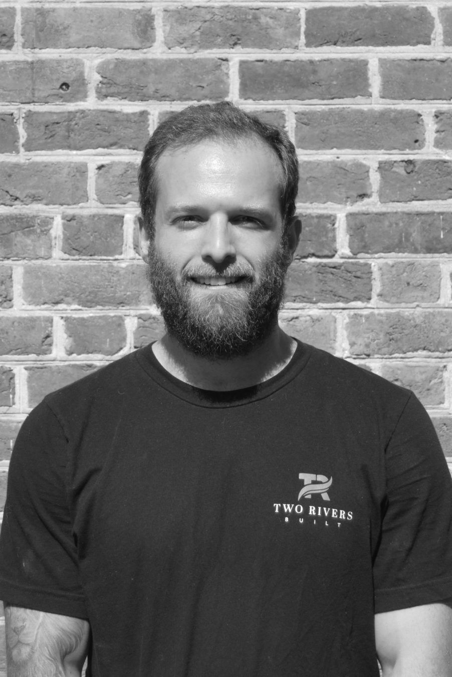 Man standing against a brick wall, wearing a dark T-shirt and smiling slightly.