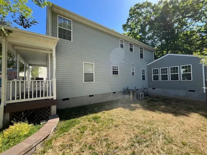 Backyard view of a light gray house with a porch, windows, and an enclosed sunroom over dry grass.